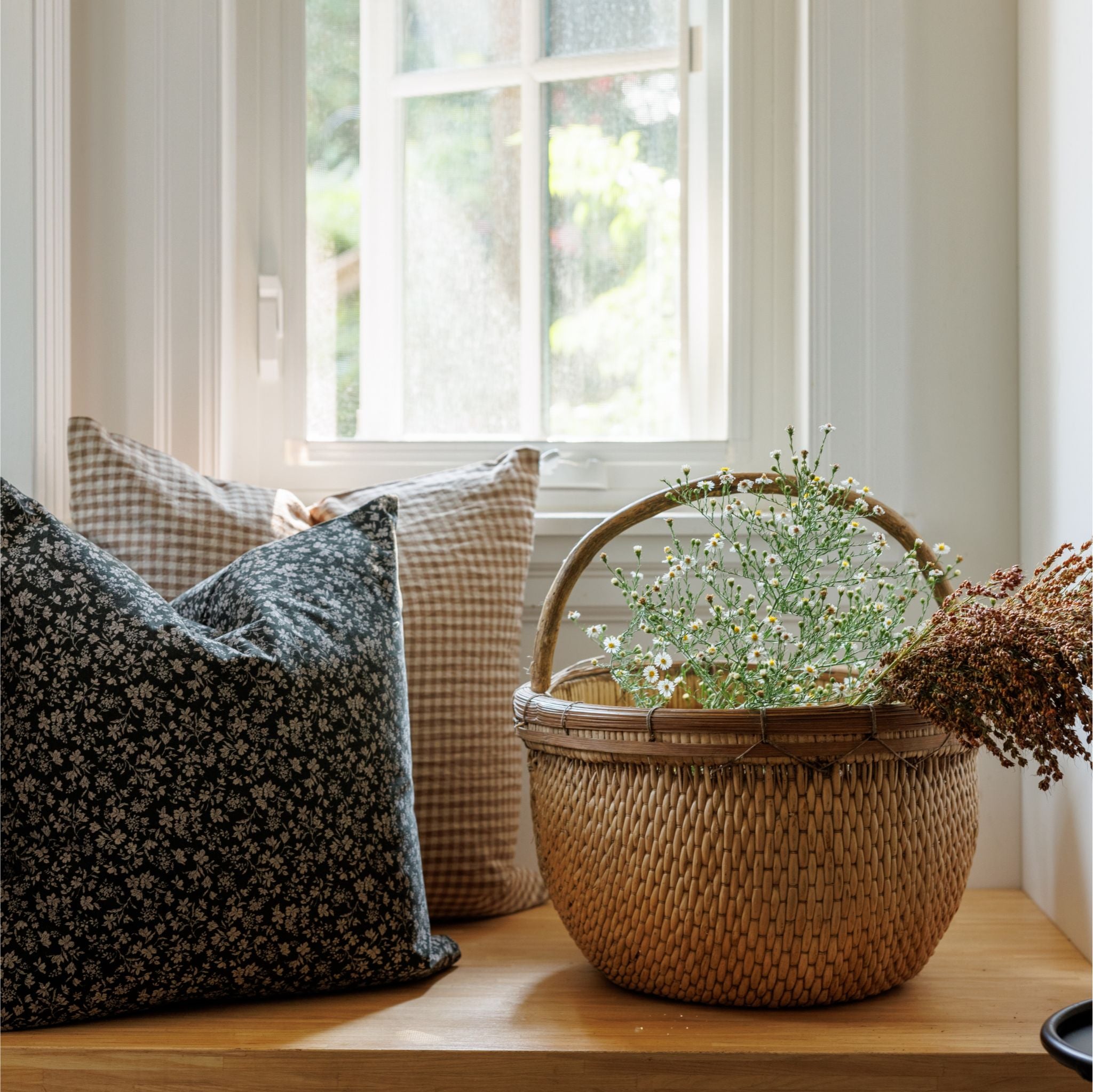 Floral pillow with brown gingham pillow on bench near window with a basket of flowers.